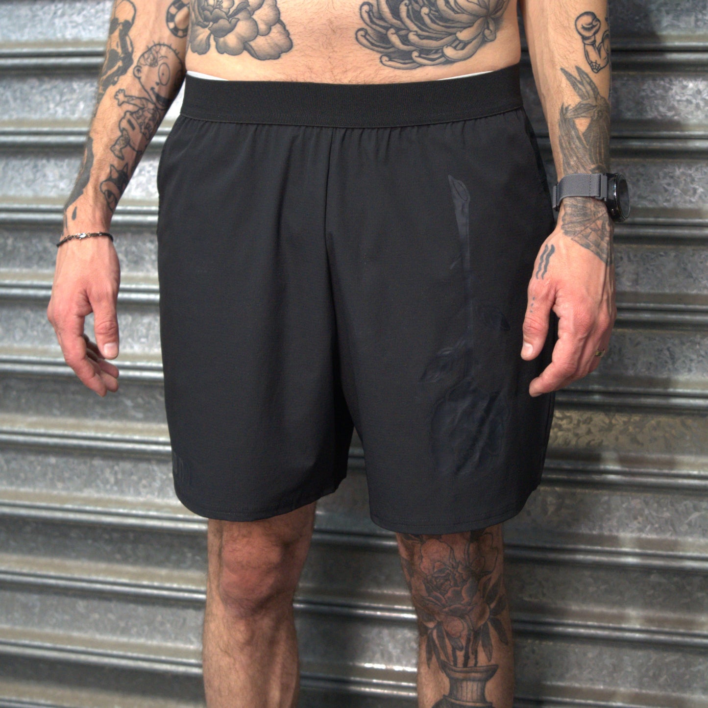 Person wearing black shorts with tattoos visible on arms and legs, standing against a metallic background.