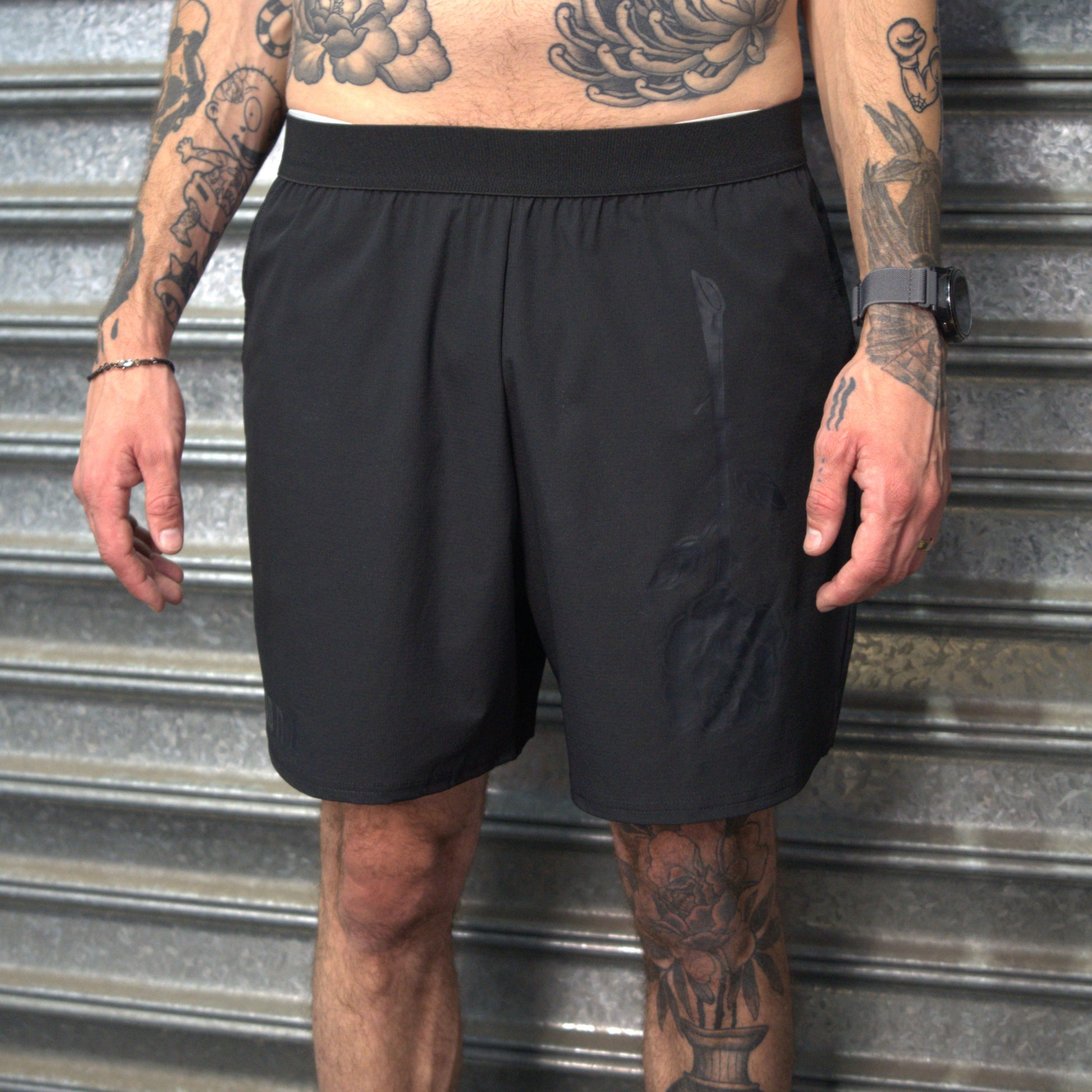 Person wearing black shorts with tattoos visible on arms and legs, standing against a metallic background.